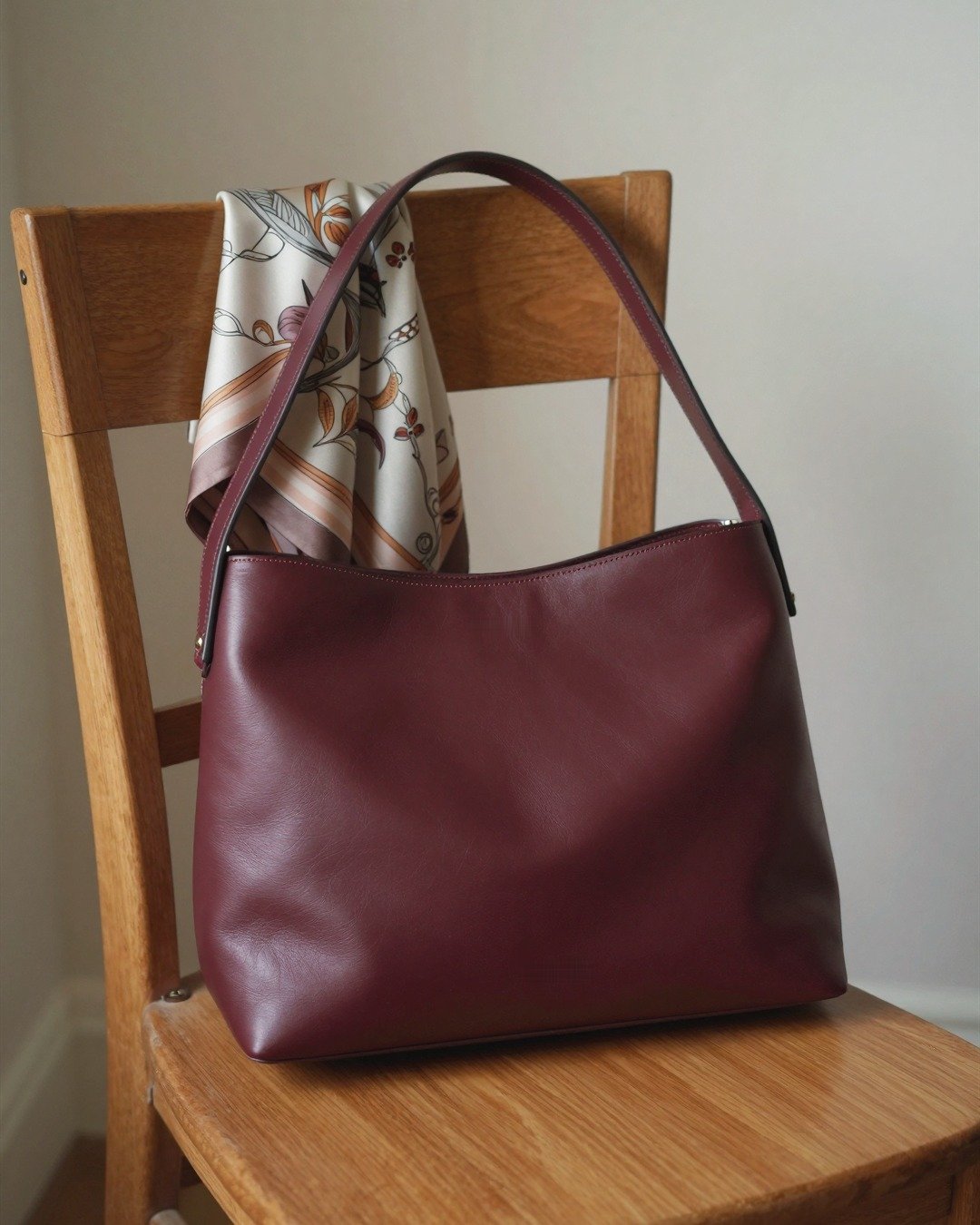 A burgundy leather bag and silk scarf on a wooden chair